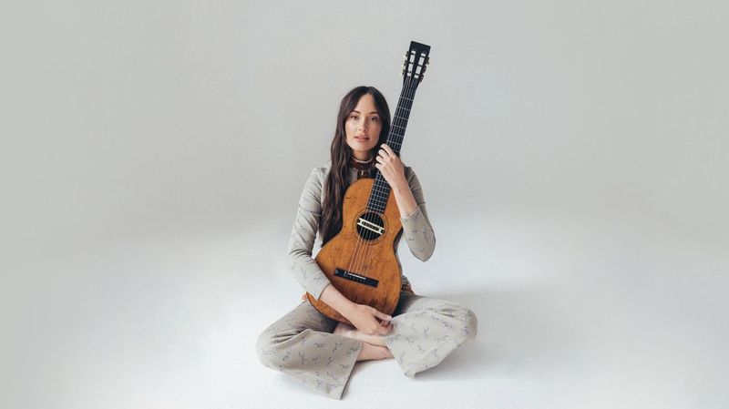 Singer Kacey Musgrave sitting on the floor and hugging a guitar against a white background.