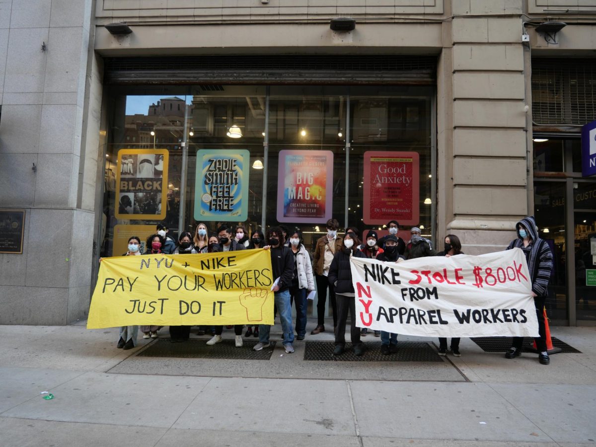 A group of people stand outside the N.Y.U. bookstore. They hold two posters, one with the words “N.Y.U. + NIKE PAY YOUR WORKERS JUST DO IT” and one with the words “NIKE STOLE $800K FROM N.Y.U. APPAREL WORKERS.”