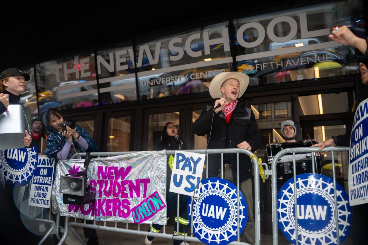 A man in a beige cowboy hat and red bandana speaks into a microphone in front of a glass building with a sign reading, “THE NEW SCHOOL.”