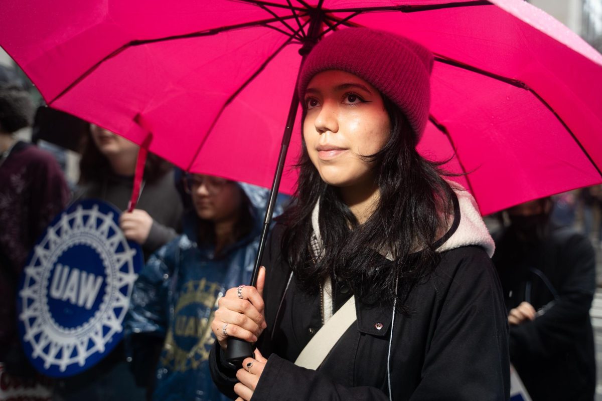 A girl with a pink umbrella watches an ongoing picket line. There is a blue U.A.W. sign in the background.