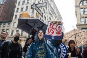 A girl in a navy blue “U.A.W.” poncho yells while holding a sign that reads, “Pay us a livin’ wage.”