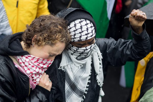 Two protestors whose faces are covered with scarves stand side by side. The protestor on the right is holding up their left fist.