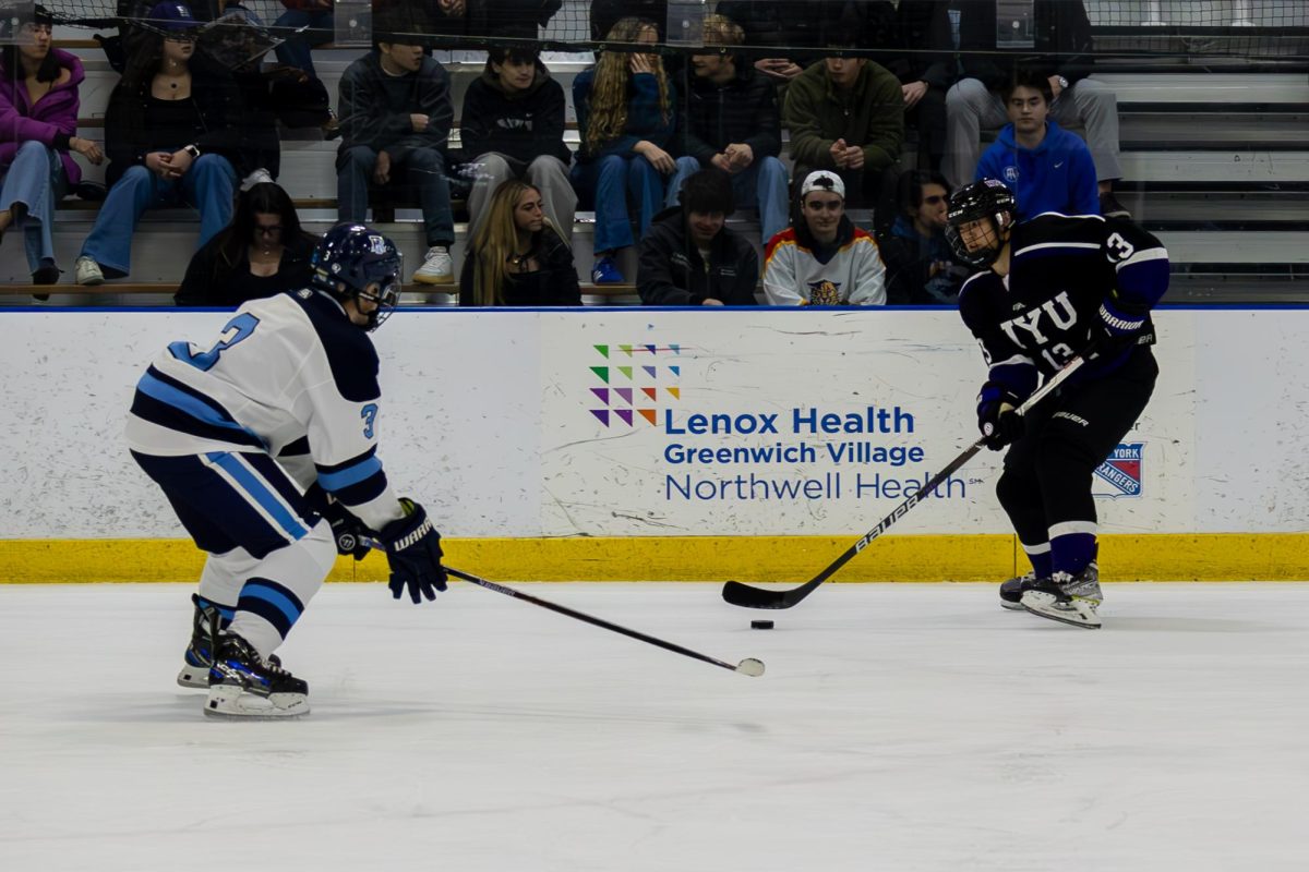 Two hockey players face each other on the ice. The right player is in a black N.Y.U. jersey and the left player is in a blue and white jersey.
