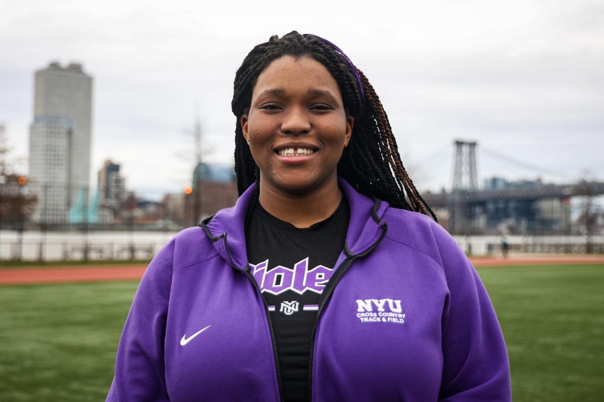 A woman with braids smiles while wearing a purple zip-up uniform with the words “N.Y.U. CROSS COUNTRY TRACK AND FIELD” in white font. Her black undershirt says “violets” in purple font.