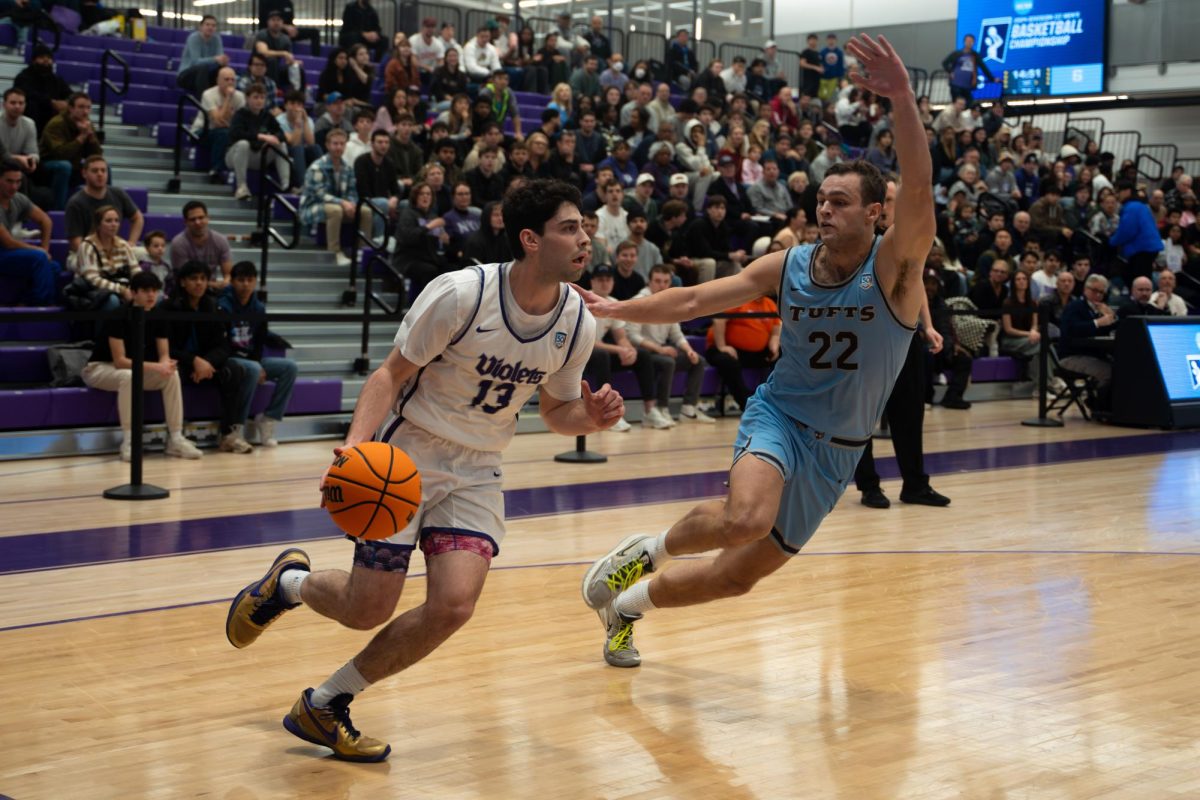 A basketball player in a white-and-purple uniform labeled “violets” runs across the court with the ball as a basketball player in a blue uniform labeled “TUFTS” attempts to block him from the right.