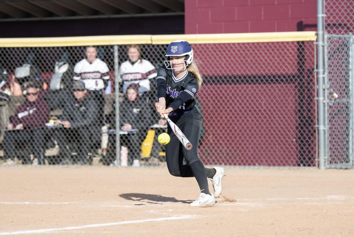 A softball player in a black-and-purple uniform in the process of hitting a ball.