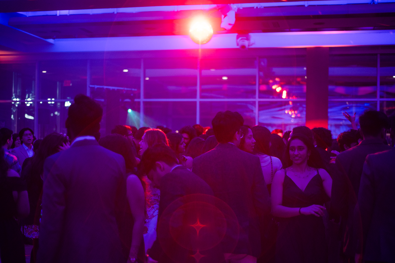 People in formalwear dance on a dance floor. The room is mostly dark, but basked in purple light. A bright orange light appears to be hanging from the ceiling.