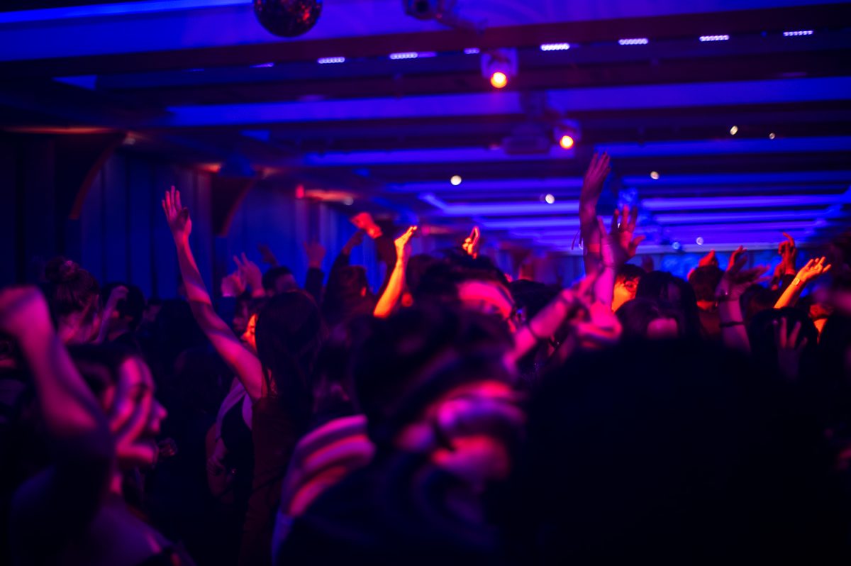 A crowd of people dance on a dance floor with their hands up. A disco ball hangs from the ceiling, and the room is basked in pink-ish purple light.