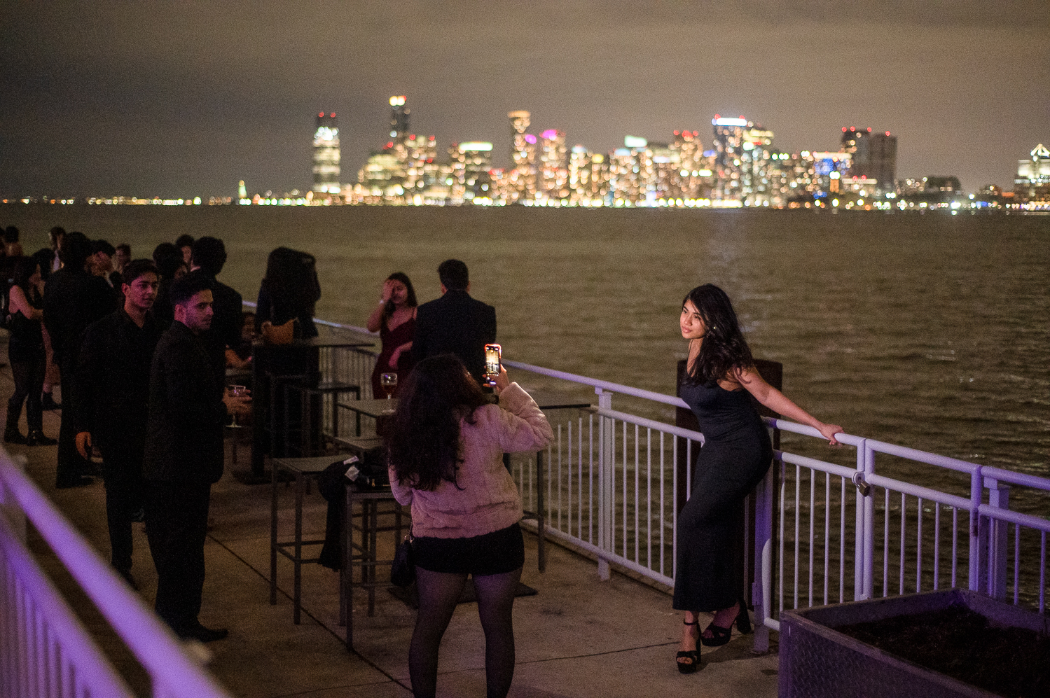 A person takes a photo of a woman in a fancy dress who is holding onto a railing behind her. Behind the subject of the photo is a large body of water, and the New York City skyline. It is nighttime, and several other people are standing in the shadows next to the pair taking the photo.