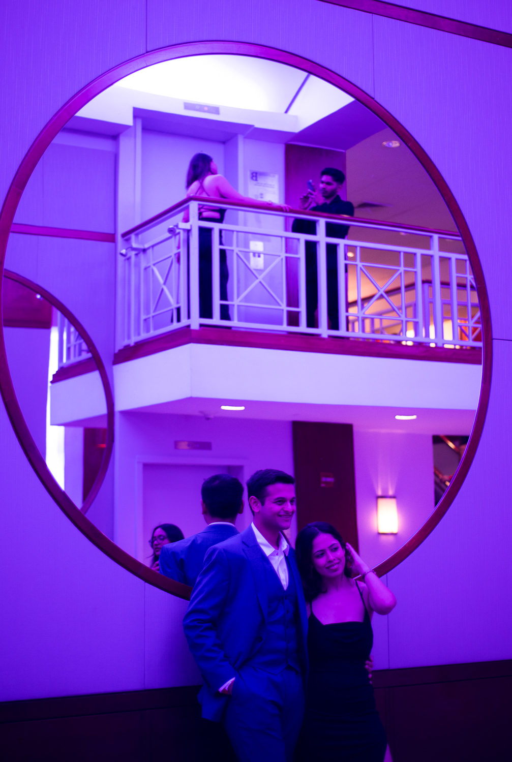 A man in a tuxedo and a woman in a dress pose side by side in front of a large, circular mirror, facing away from it. Through the mirror, the back of a woman leaning against a balcony railing corner, posing for a photo being taken by a man, is visible. The room is basked in blue-ish purple light.