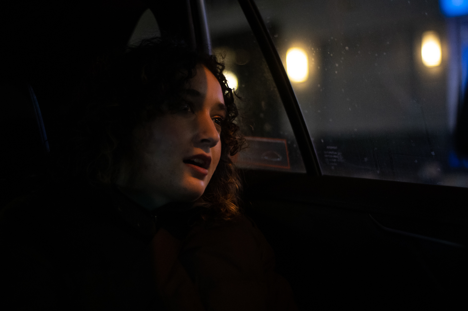 A woman with curly hair and half her face dulled by shadows sits at the window seat in a car. It is nighttime outside the window.