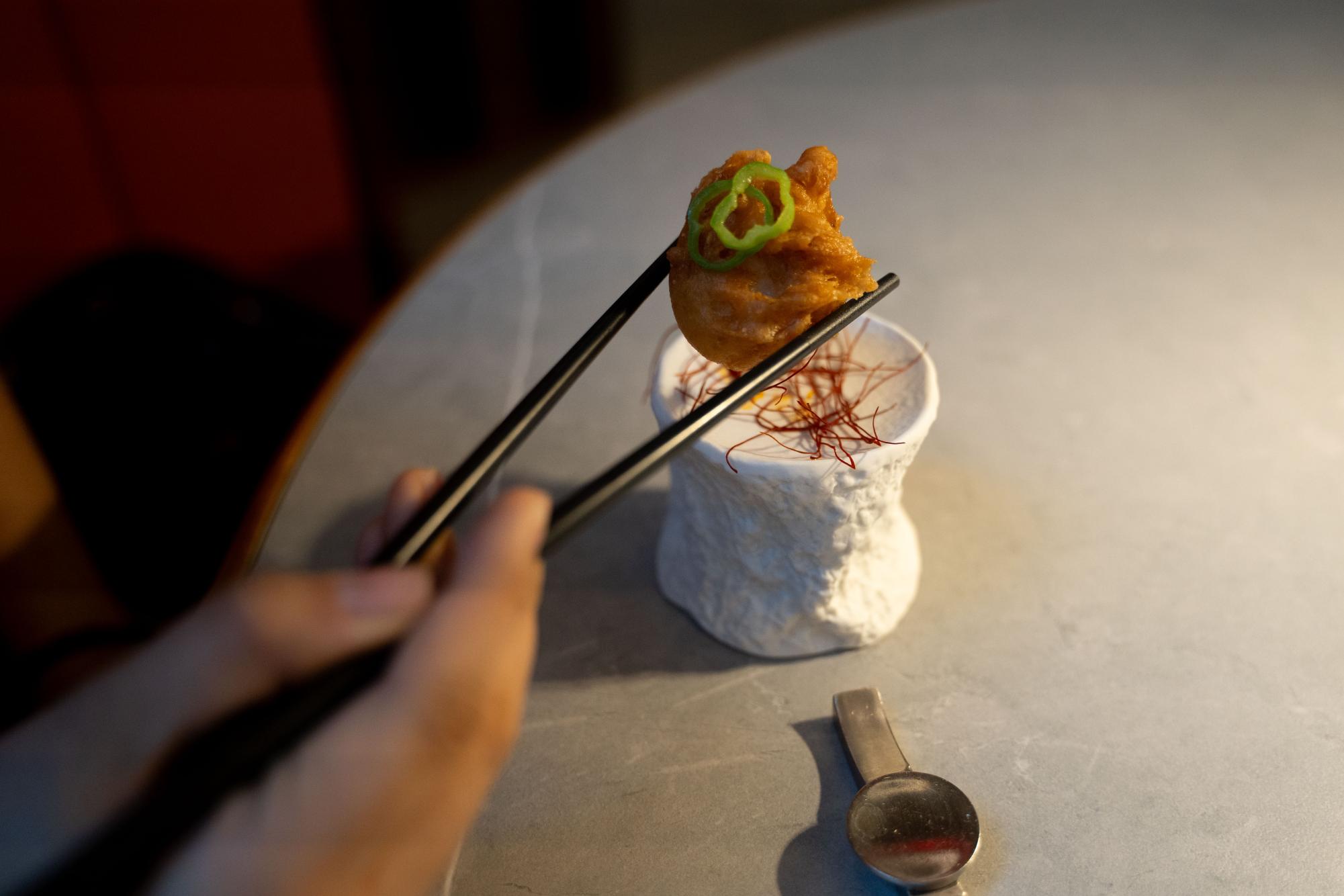 A hand holding chopsticks picks up a small piece of fried scallop garnished with pepper slices on an artisanal stand.