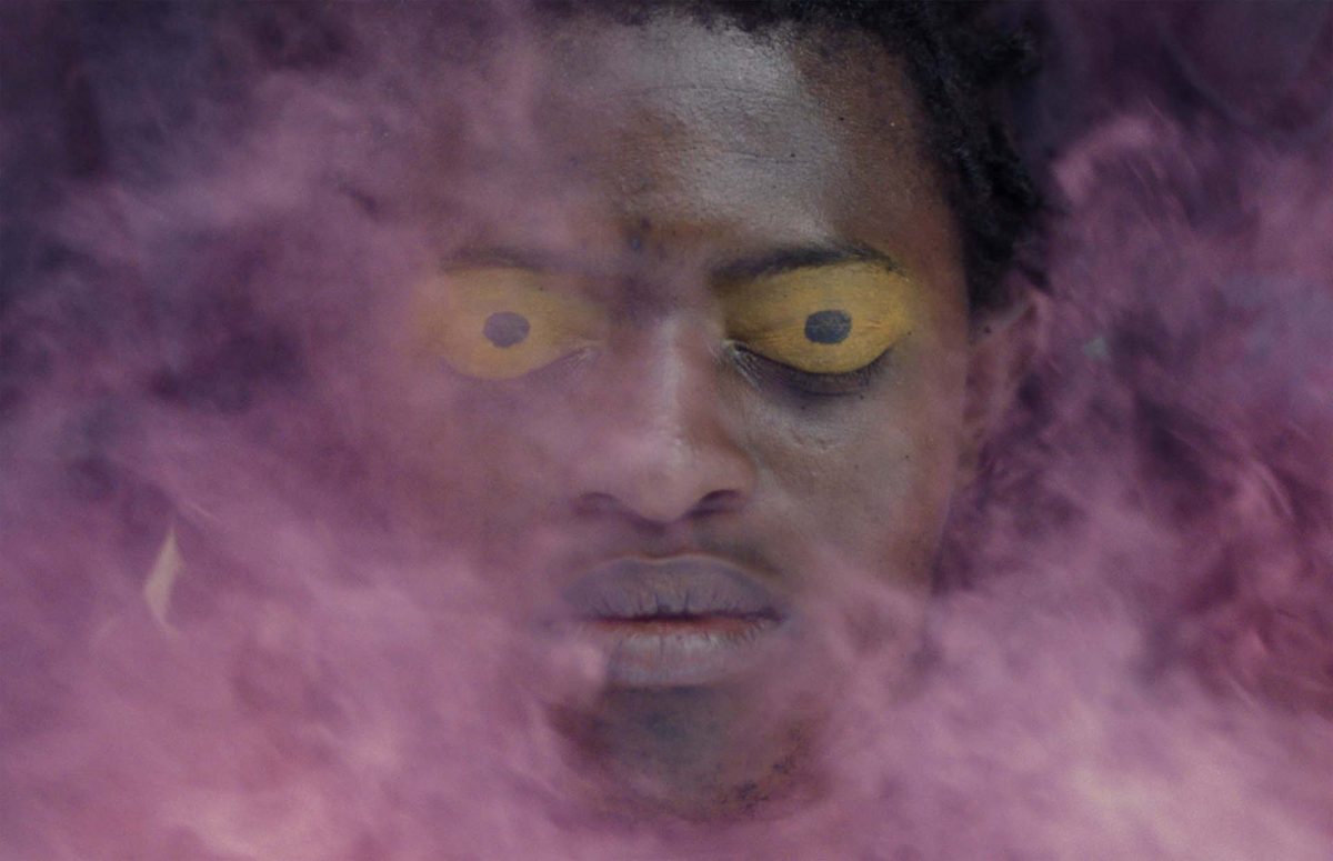A photograph of a man’s face with his eyes closed and his eyelids are painted yellow with a black dot in the middle. There is pink smoke in front of his face.