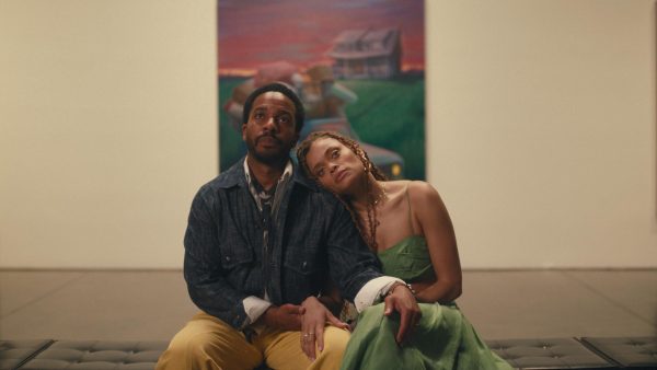 Two people sitting on a bench in a museum, looking at an out-of-frame painting. The woman is leaning on the man, and they are holding hands.