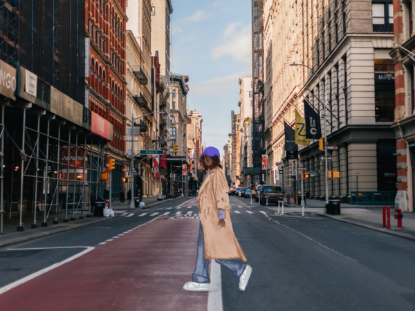 A person crossing a city street is wearing a tan trench coat, purple baseball cap, blue jeans and white sneakers.