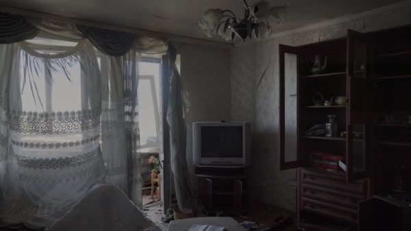 A dark, messy room with an open balcony on the left, a TV in the middle, and an open armoire on the right. The wallpaper is peeling, a window is broken and there is glass on the floor.