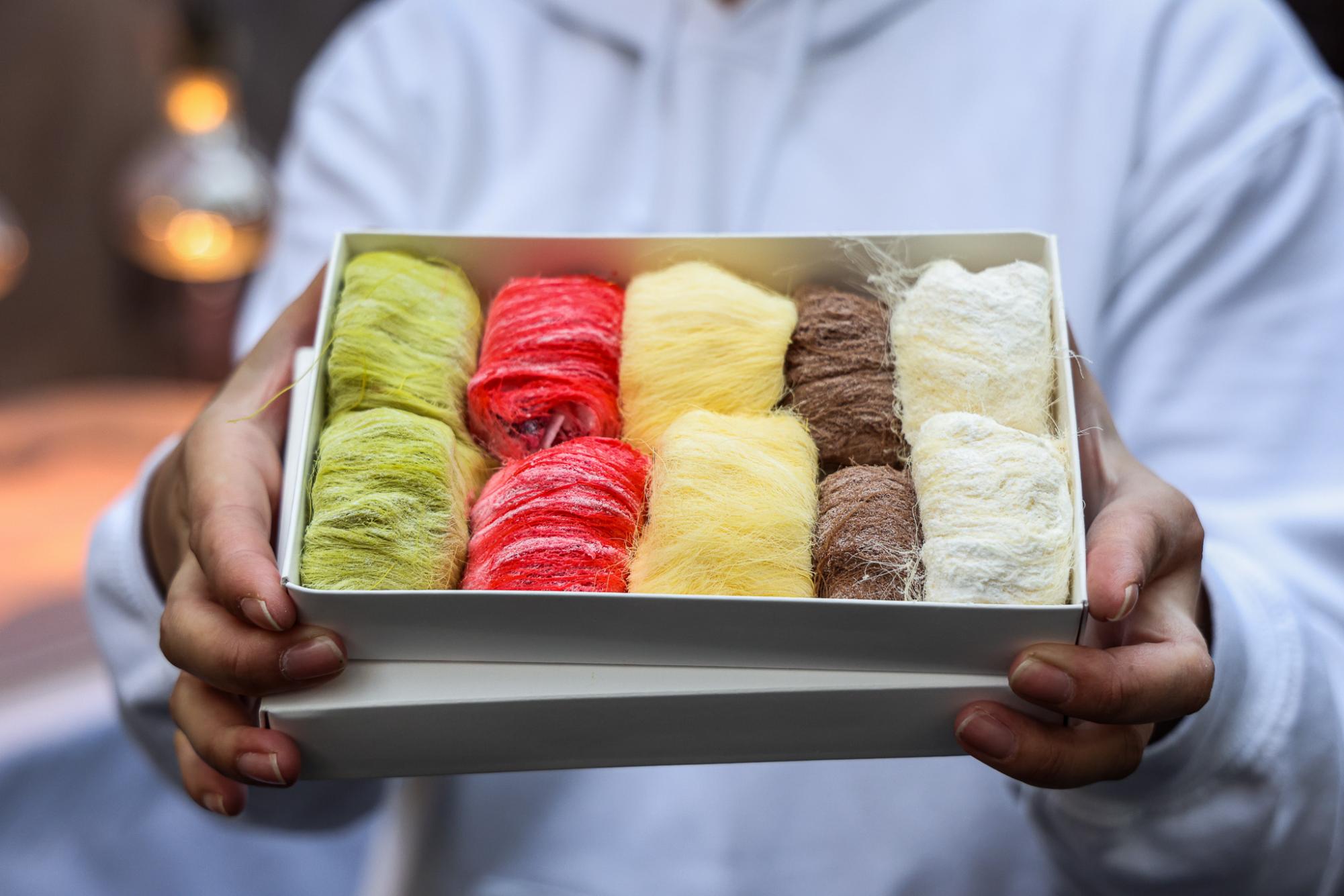 Person holds out a decorative box filled with an arrangement of green, red, yellow, brown and white string candy.