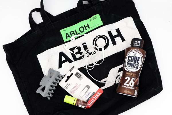 A black tote bag lays flat on a white surface. On top of it are a gray claw clip, a flash drive, a lip oil, a pair of wired earbuds and a protein shake.