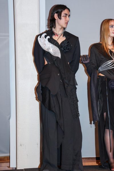 A model wears a black draped suit with a black rose and a silver metal accent.