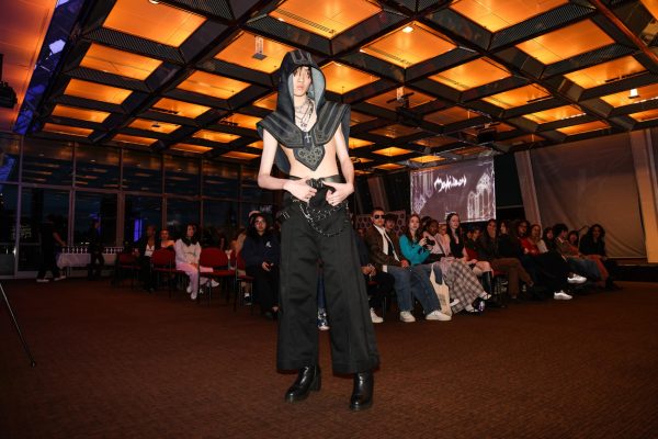 A model poses wearing a headpiece with connected shoulder pads, black slacks and heeled boots.