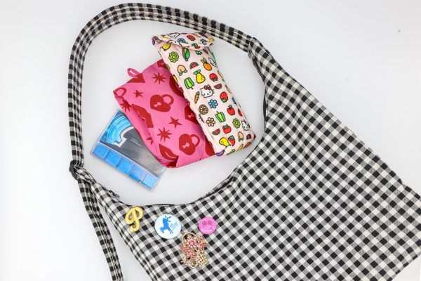 A black-and-white checkered bag lays flat on a white surface. On the bag are four assorted pins. Beside the bag are a pencil pouch with small cartoon symbols, a pink pouch with red symbols and a pack of gum.