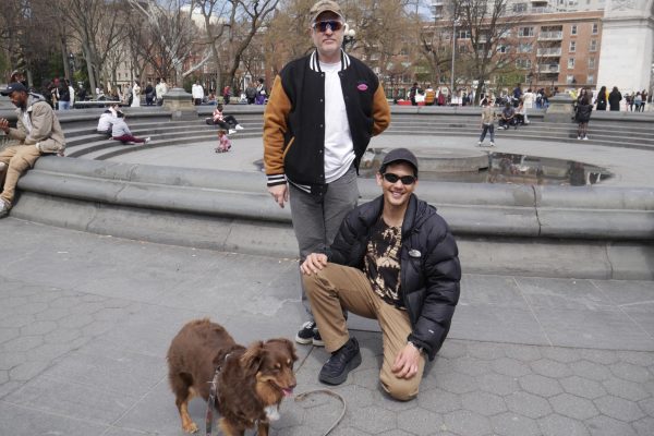 Two men posing with a dog at a park. One man stands in a black and gold jacket, white shirt, and black pants while the other squats in khaki pants and a black puffer, next to the medium-sized brown dog.