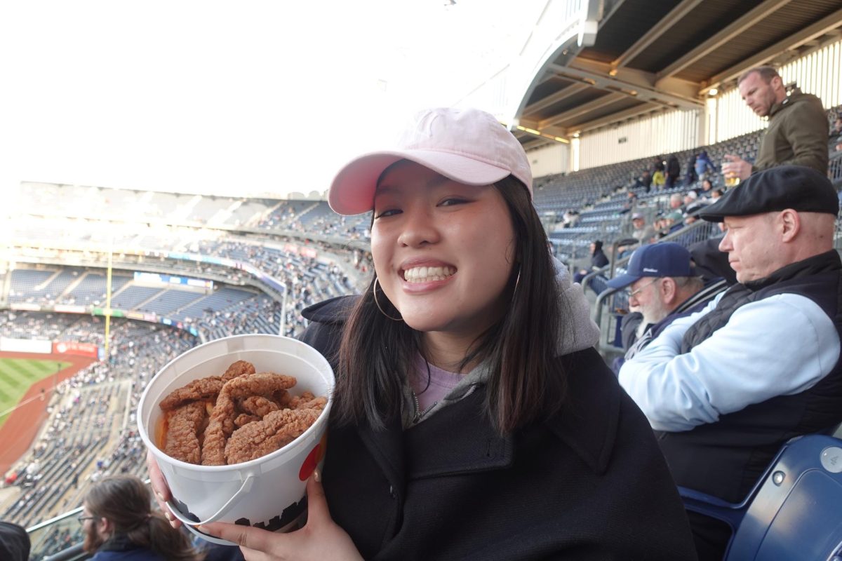 A girl sitting in a baseball stadium smiles while holding a bucket of fried chicken.
