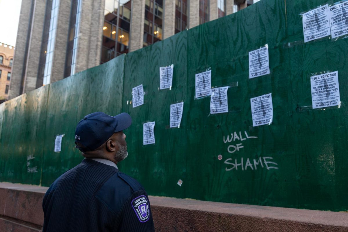 A campus safety officer looks at a green wall which has posters and the words “WALL OF SHAME” on it.