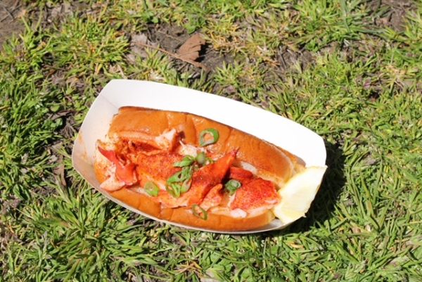 A lobster sandwich with scallions on top in a white container sitting on grass. 