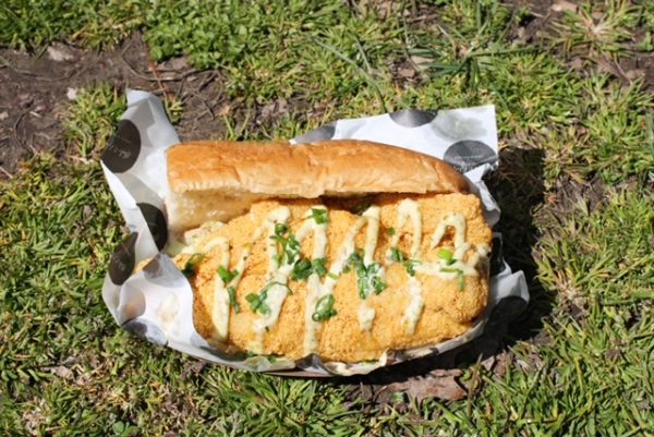 A fish sandwich with dressing and scallions on top laying on a paper on the grass. 