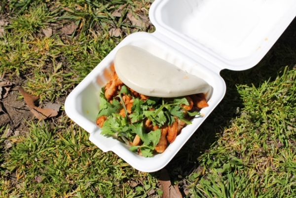 A white bao bun containing pieces of pork and cilantro in a white container sitting on grass. 