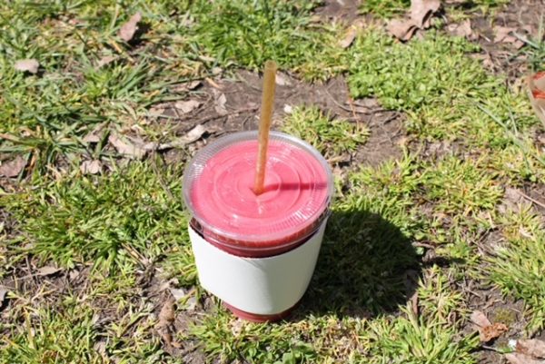 A pink smoothie with a straw and white cup holder sitting on grass. 