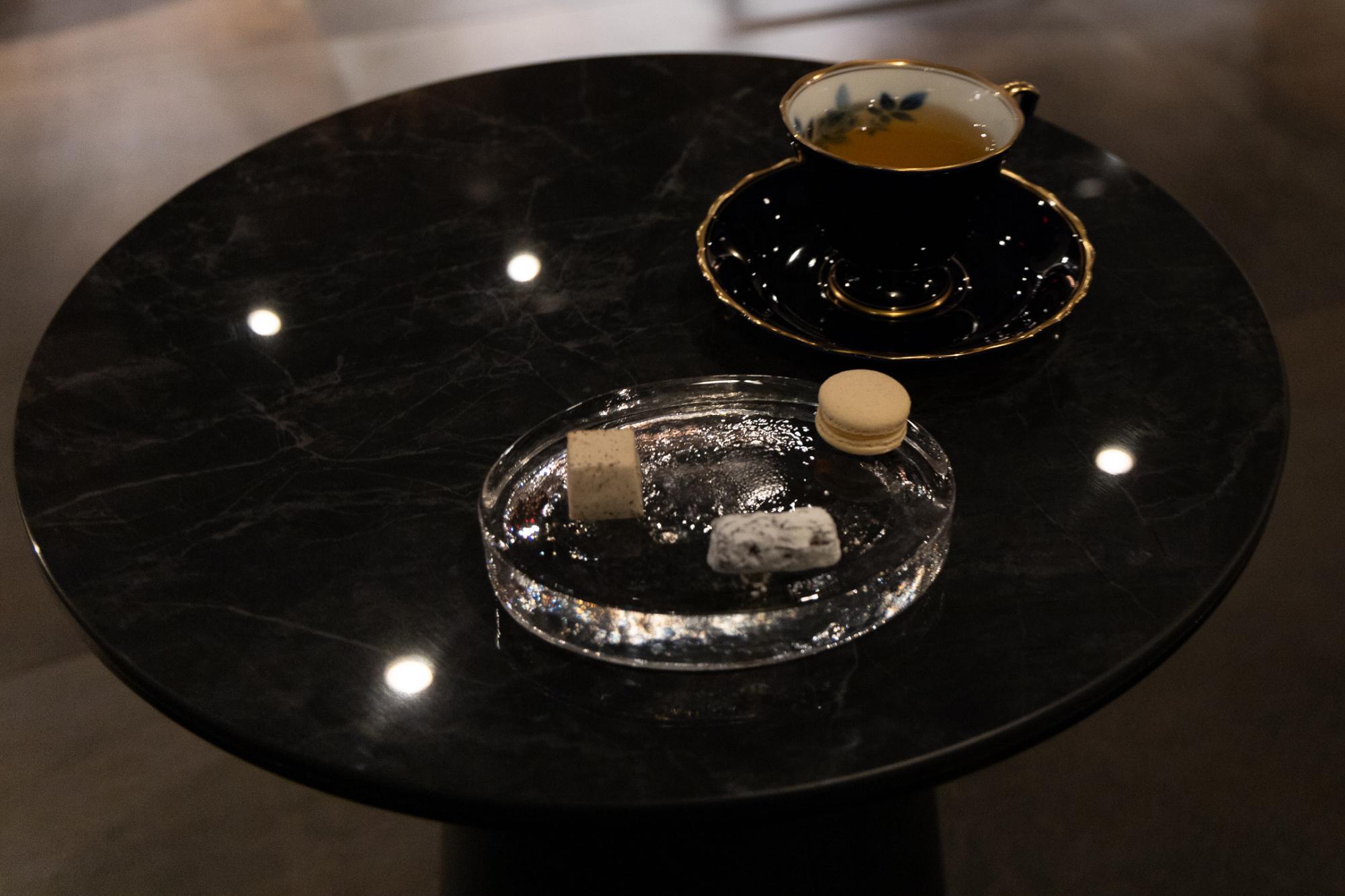 A teacup filled with yellow tea and three small desserts on a black table.