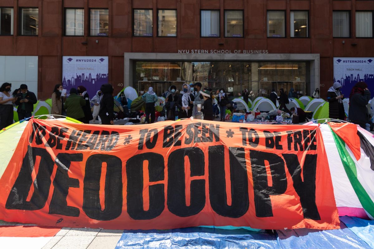 A red banner that has the words “TO BE HEARD TO BE SEEN TO BE FREE” above the word “DEOCCUPY.” In the background protestors stand around tents in front of a building that is labeled “NYU STERN SCHOOL OF BUSINESS.”