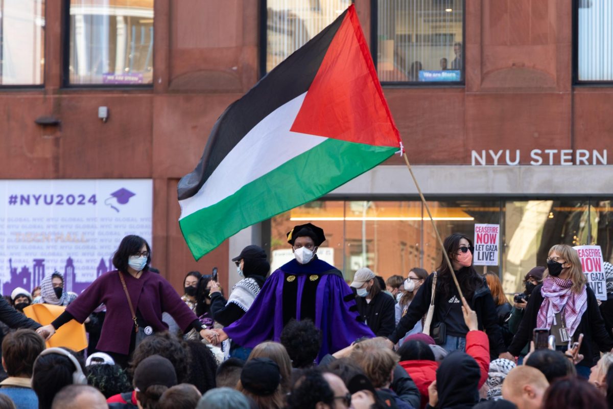 Protesters hold their hands in front of a building that is labeled “N.Y.U. STERN” as a Palestinian flag waves overhead.