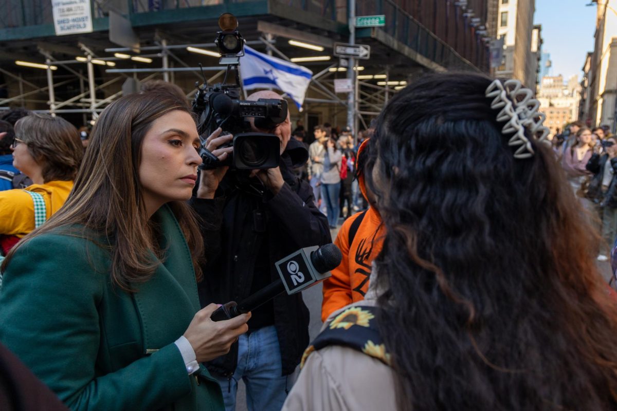 A woman in a green coat holds a microphone, interviewing a student at a protest.