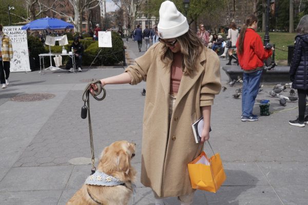 A woman with a long, beige coat, brown hair, and white hat holds out her large, blonde, fluffy dog’s leash.