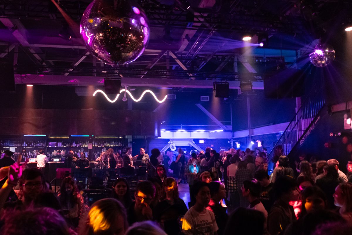 A nightclub with purple-blue lighting full of people. From the ceiling hangs a disco ball.