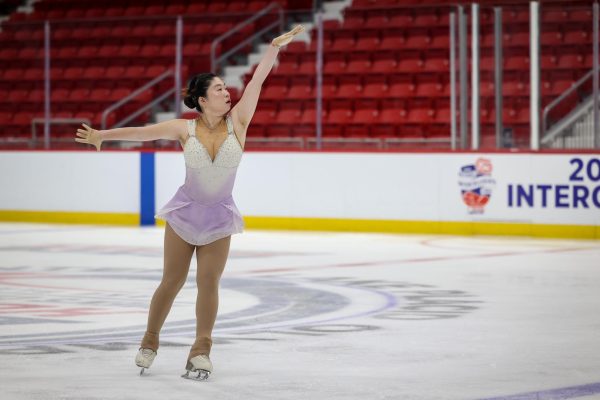 A figure skater on the ice with their left arm raised up and their right arm straight.
