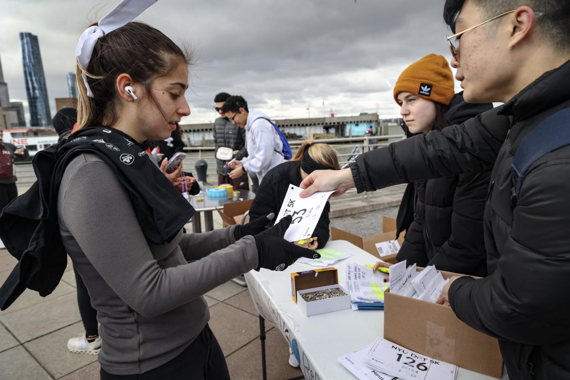 A man hands a participant her running number in front of the New York City shoreline.