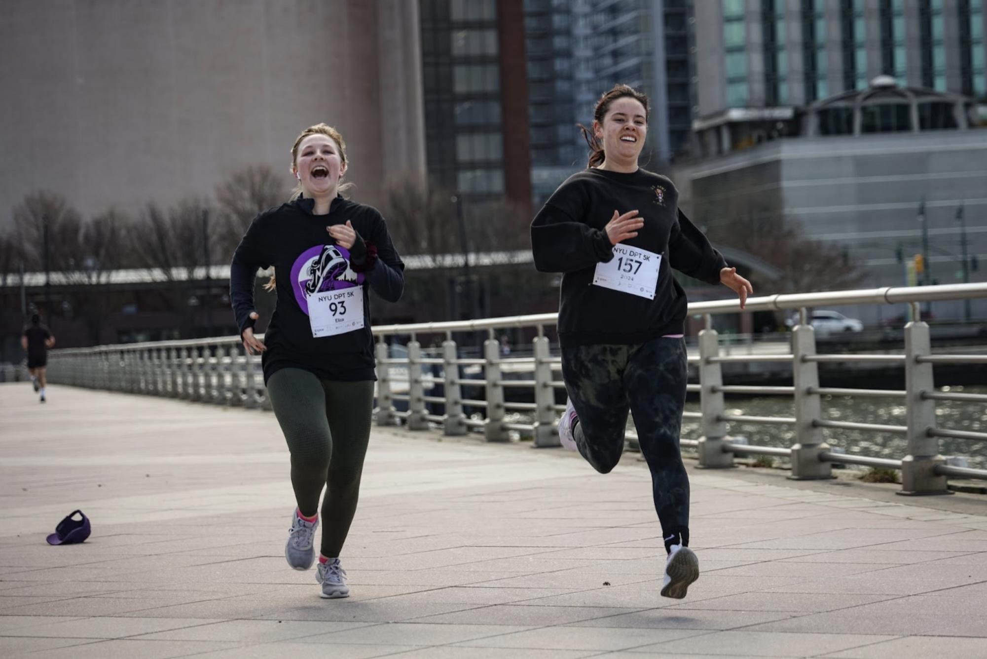 Two women with running bibs pinned to their shirts run on a bridge.