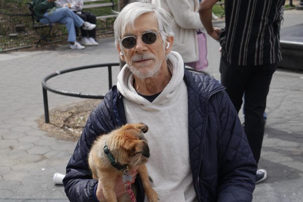 A man with white hair, a white hoodie, blue puffer and sunglasses holds a small beige dog with a green collar.