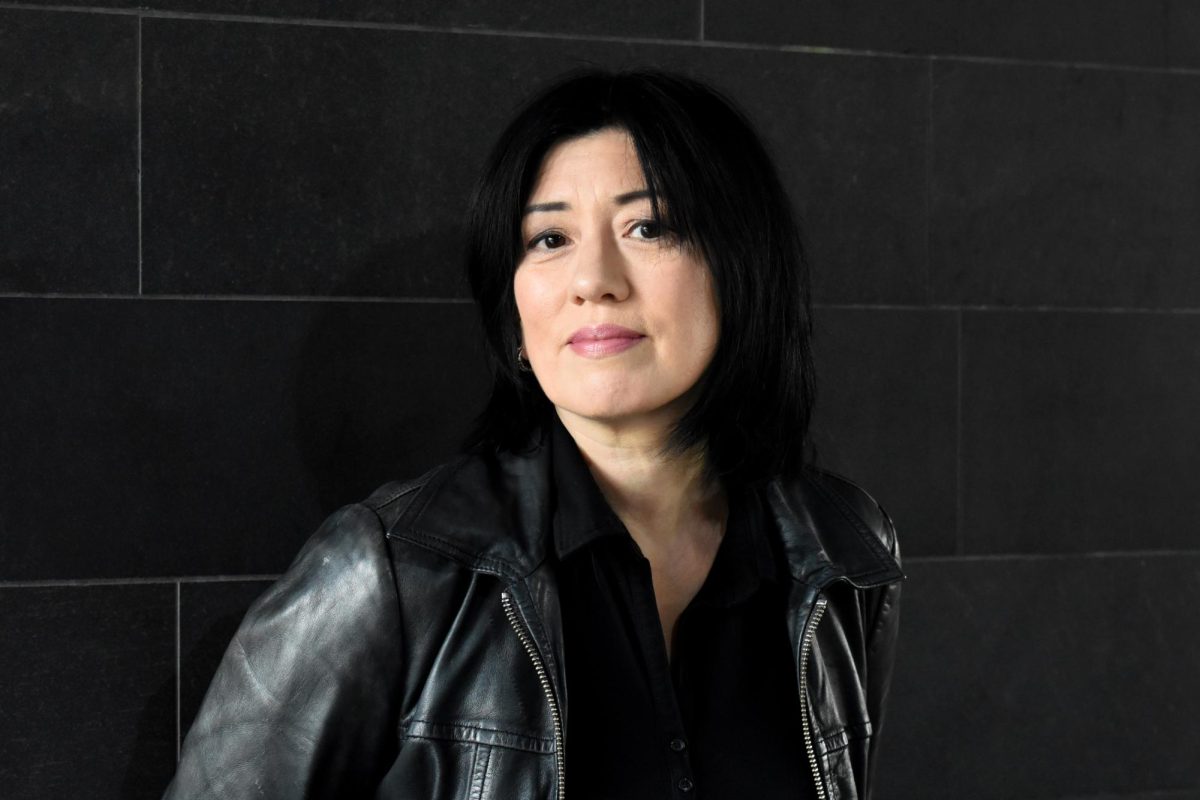 A woman with black hair, wearing a black shirt and black leather jacket, stands in front of a gray tiled wall.