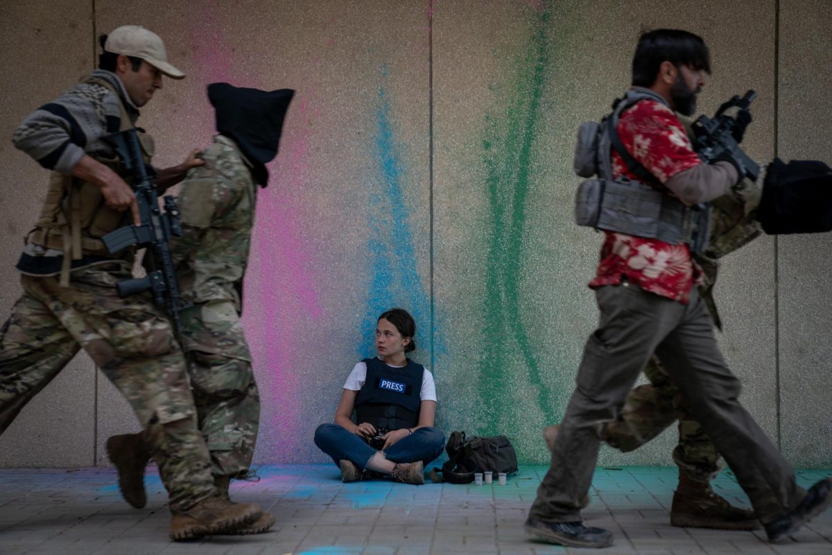 A film still of two men in military outfits holding guns and walking people with black bags over their heads. Behind them, a girl sits against a wall with chalk markings of pink, blue and green, wearing a black vest with the word, “PRESS” written on it.
