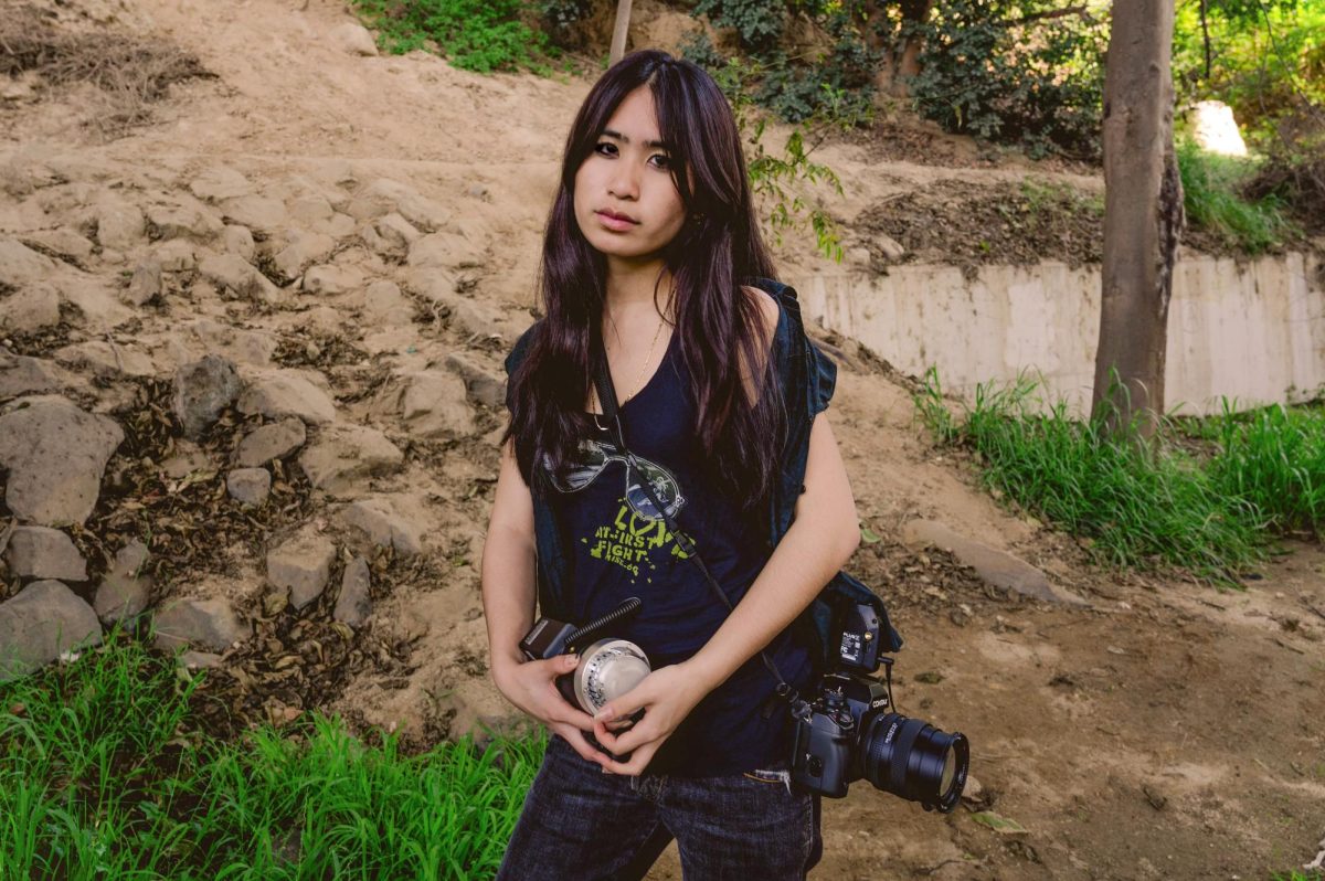 A girl wearing a black shirt holds a camera at her side while posing in a sandy outdoor area.