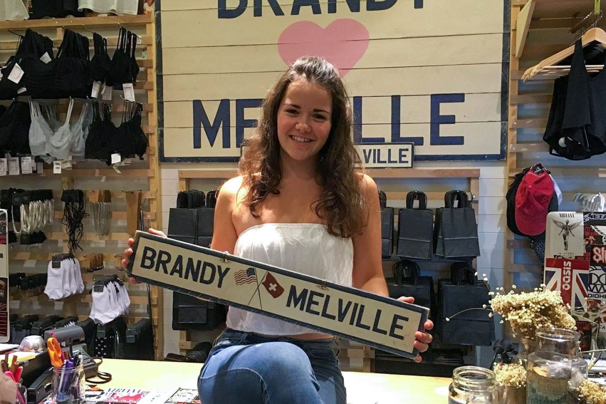 A+girl+wearing+a+white+tank+top+and+blue+jeans+sits+on+a+store+countertop%2C+holding+a+wooden+sign+that+reads%2C+%E2%80%9CBrandy+Melville.%E2%80%9D