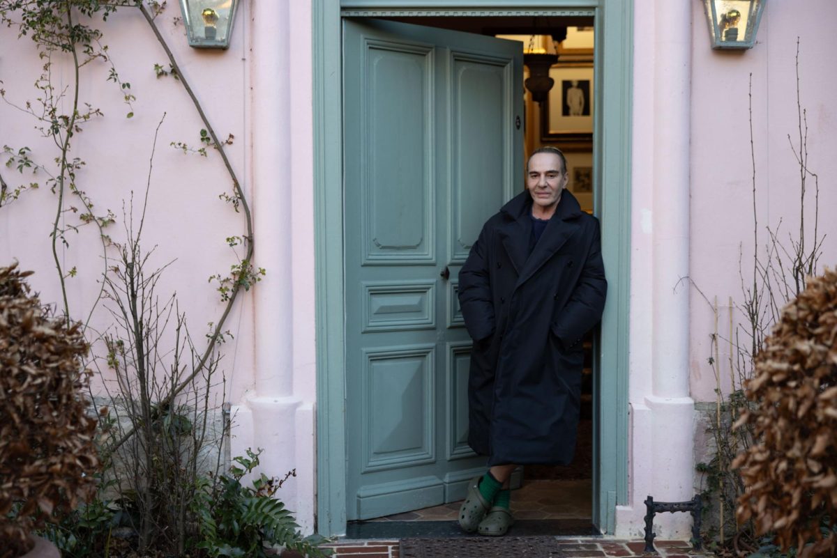 A man wearing a black trench coat and green crocs leans on a teal front door.