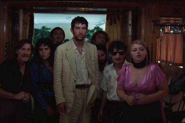 Movie screengrab of five people gathered in a room entrance, the central figure is a brunette man in a tan suit.