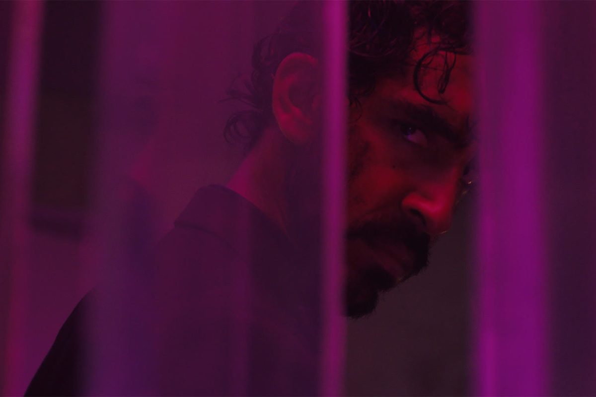 A sweaty man with blood on his face glares through a curtain in purple lighting.