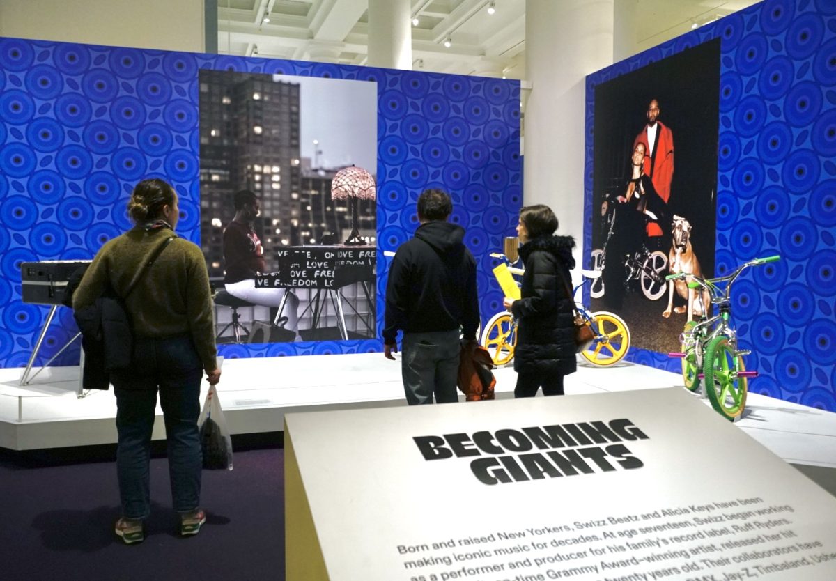 Three people around a museum exhibit with blue walls displaying a photo of a person sitting at a graffitied piano and a photo of a woman, a man and a dog with a bike. Two neon bikes and a piano are on a raised platform in front of the photos.